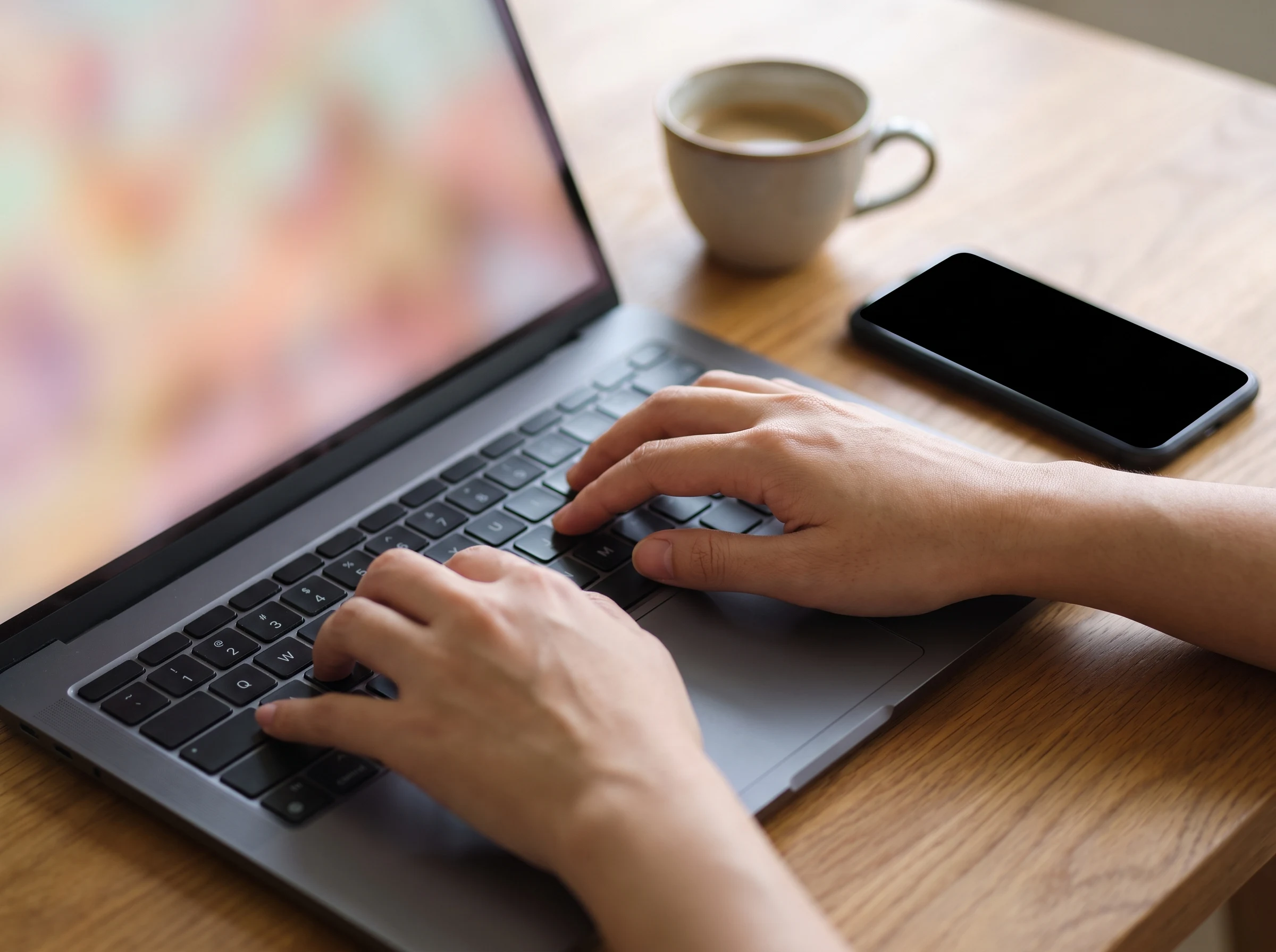 Close-up of hands typing on a laptop with a blurred colourful screen, coffee and a blank phone