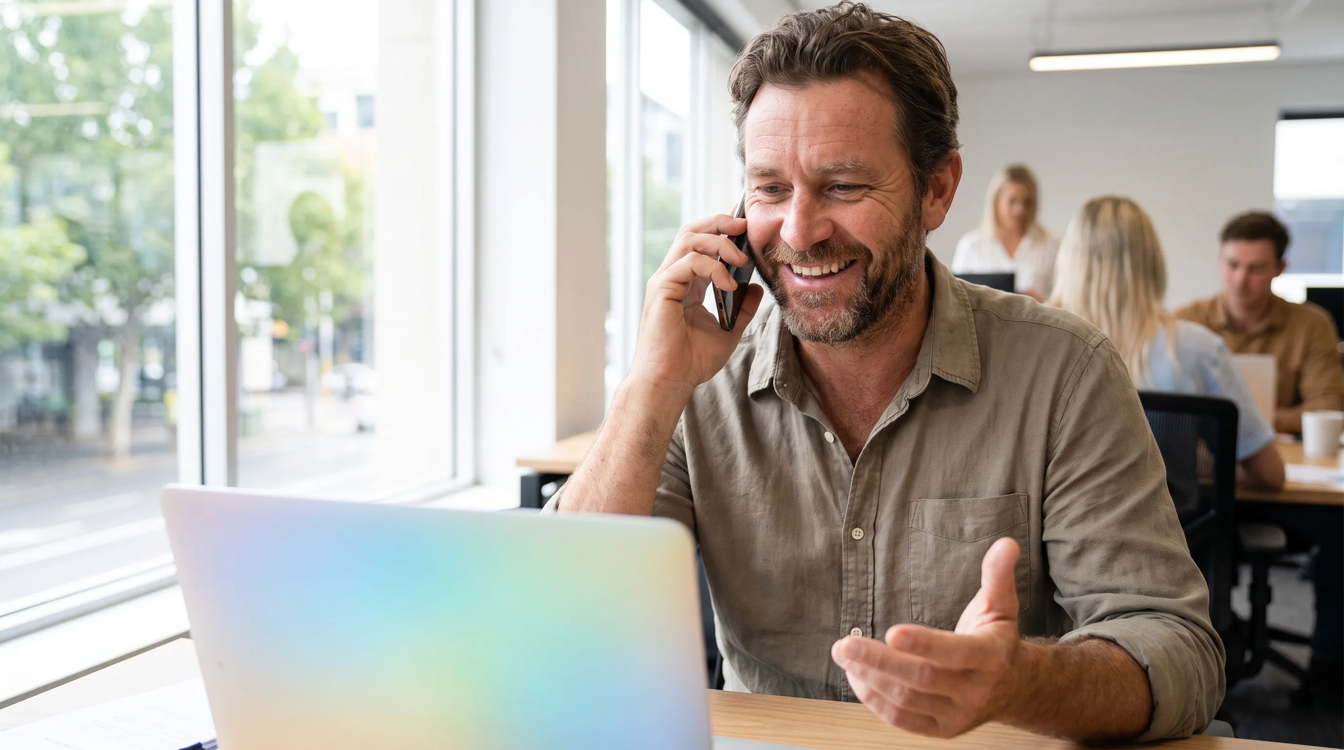 Friendly Australian professional smiling on the phone while using a laptop in a bright office